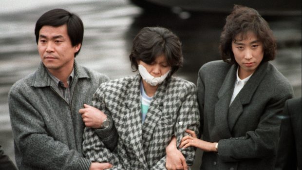 Jihokorejští vyšetřovatelé svírají paže severokorejské agentky Kim Hjon-hi, která spolu se svým kolegou Kim Song-ilem provedla teroristický útok na letadlo Korejských aerolinek. Bomba, kterou maskovaný pár nastražil na palubě letadla, zabila 115 lidí. Tento známý snímek vznikl 15. prosince 1987, šestnáct dnů po útoku (29. listopadu 1987).