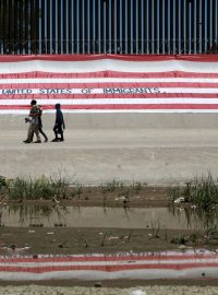 Migranti procházejí kolem velké americké vlajky, která pokrývá část mexicko-americké hranice