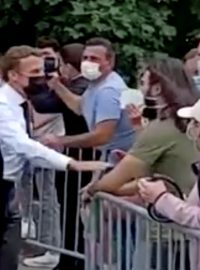 Prezident Emmanuel Macron (v bílé košili) se přiblížil k davu lidí za mobilní bariérou, zamířil k muži s bílou rouškou, zeleným trikem a dlouhými vlasy, chytil ho za předloktí a on mu dal facku